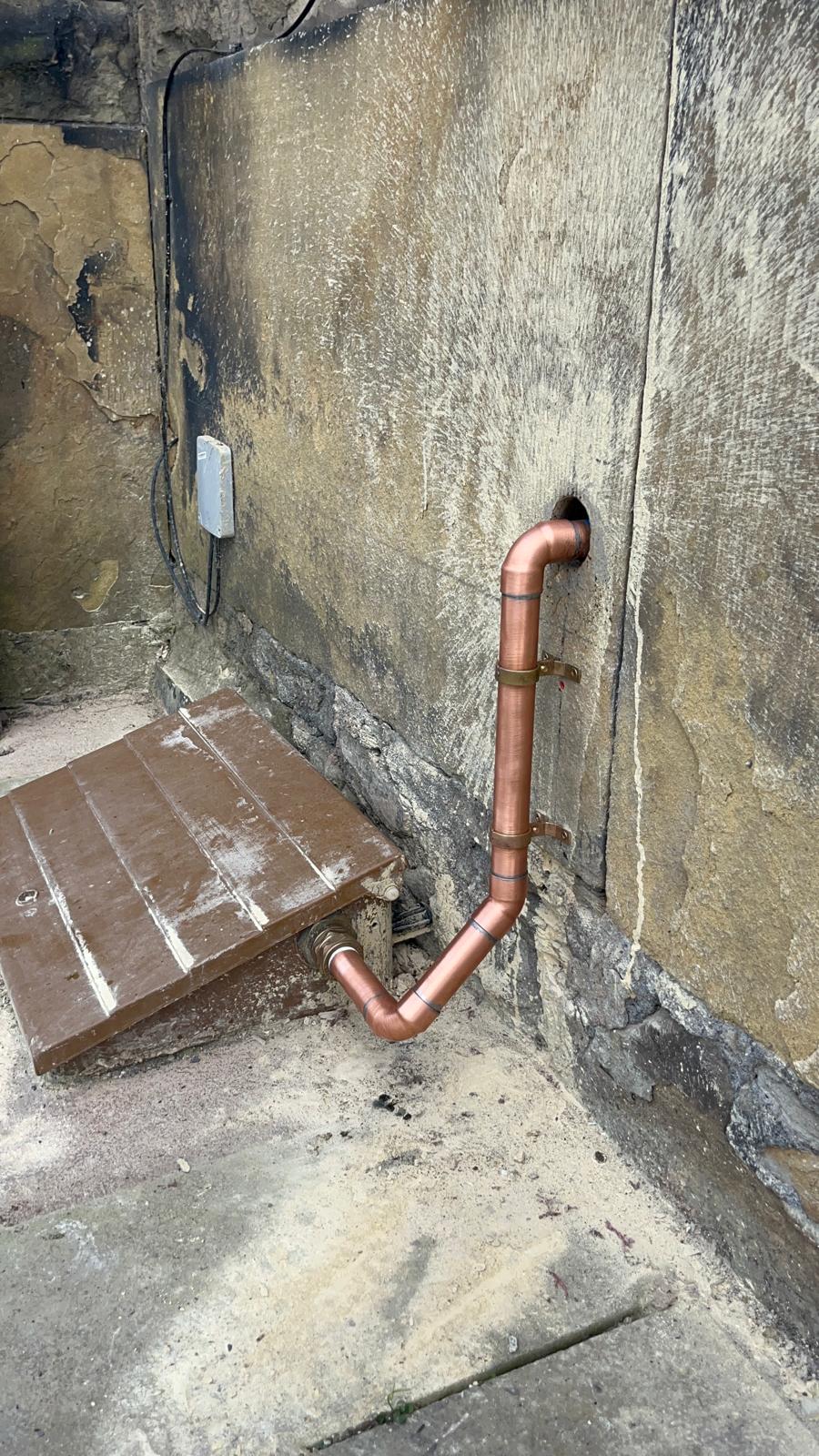 Neatly clipped copper pipe running down an exterior stone wall to a drain connection
